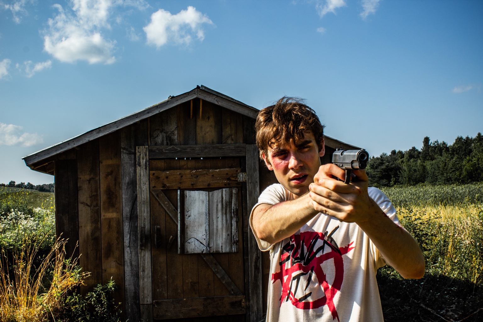An injured young man aims a handgun in front of the shed